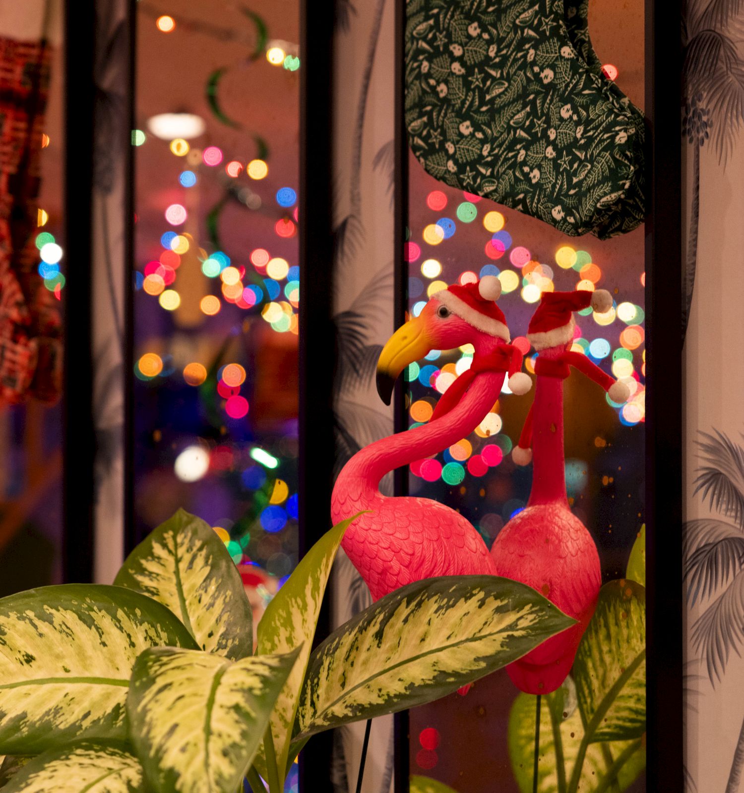Vibrant holiday scene with stockings, colorful lights, flamingo decorations, and lush plants create a festive atmosphere indoors.