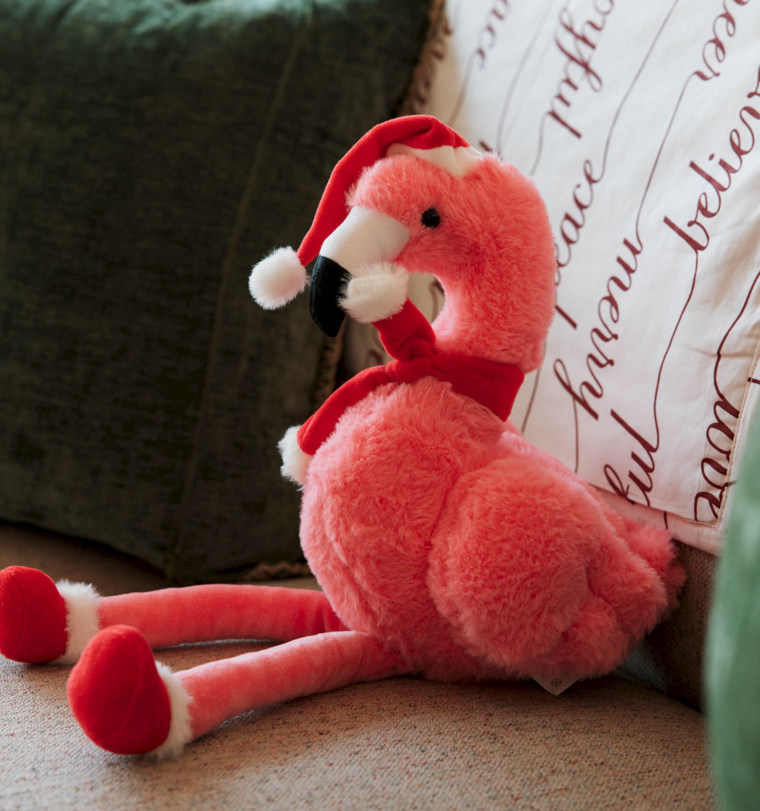 A plush pink flamingo toy wearing a Santa hat and scarf sits on a sofa, surrounded by green and white cushions with script print.