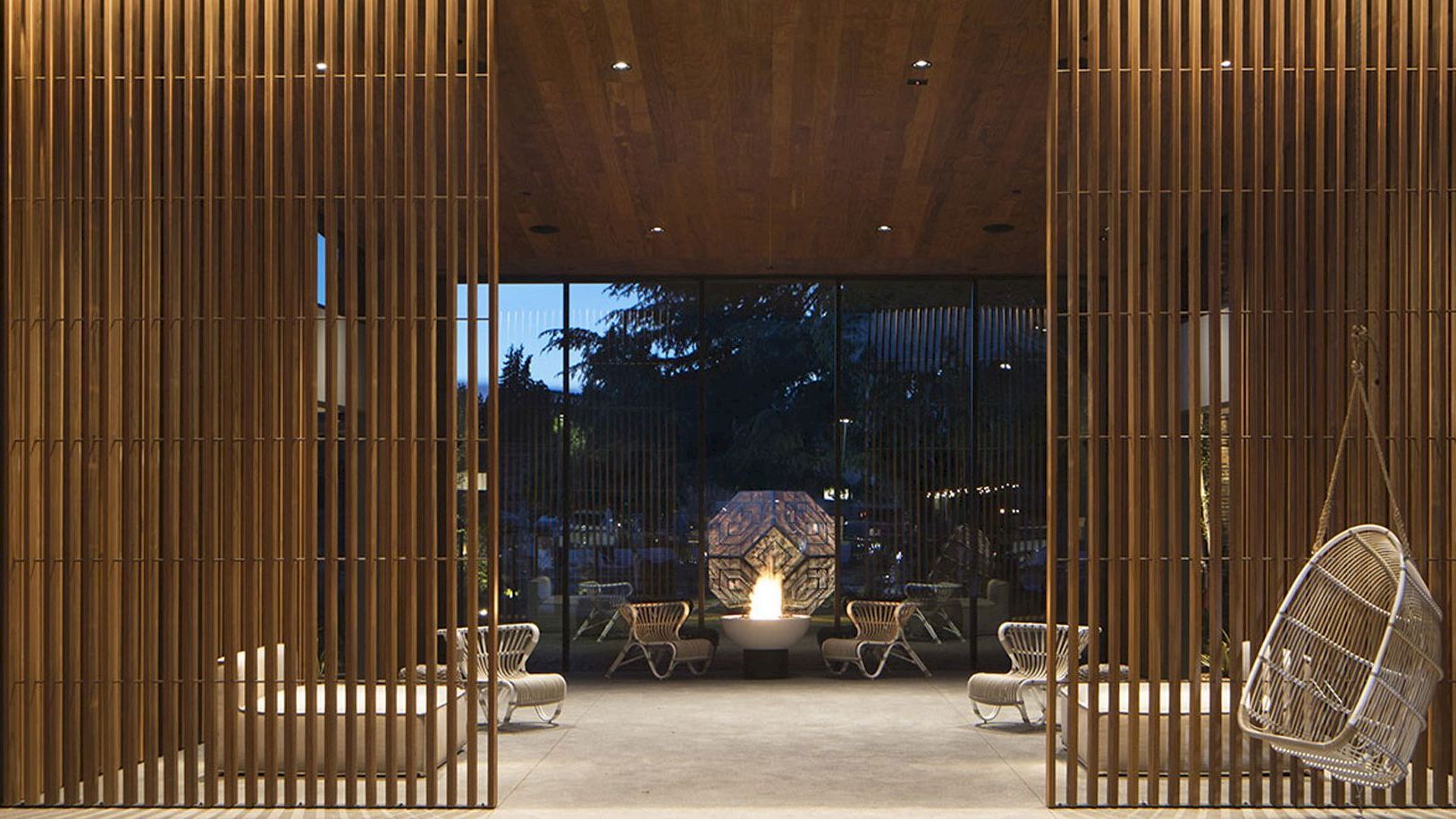 A modern, open-space lobby with wooden slats, hanging chairs, and outdoor seating areas arranged around a central fireplace, seen from outside.