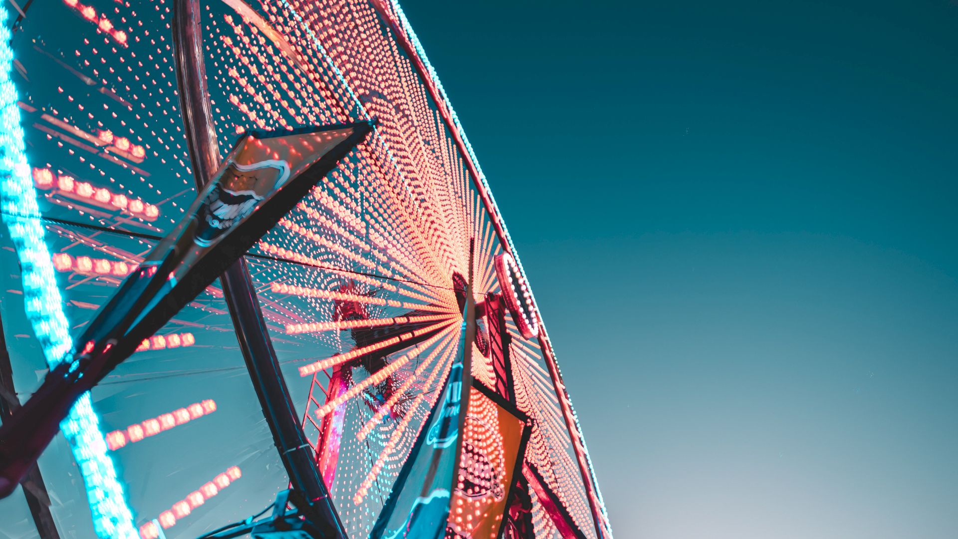 The image shows a close-up of a brightly lit Ferris wheel against a clear sky, with colorful lights creating a vibrant and dynamic pattern.