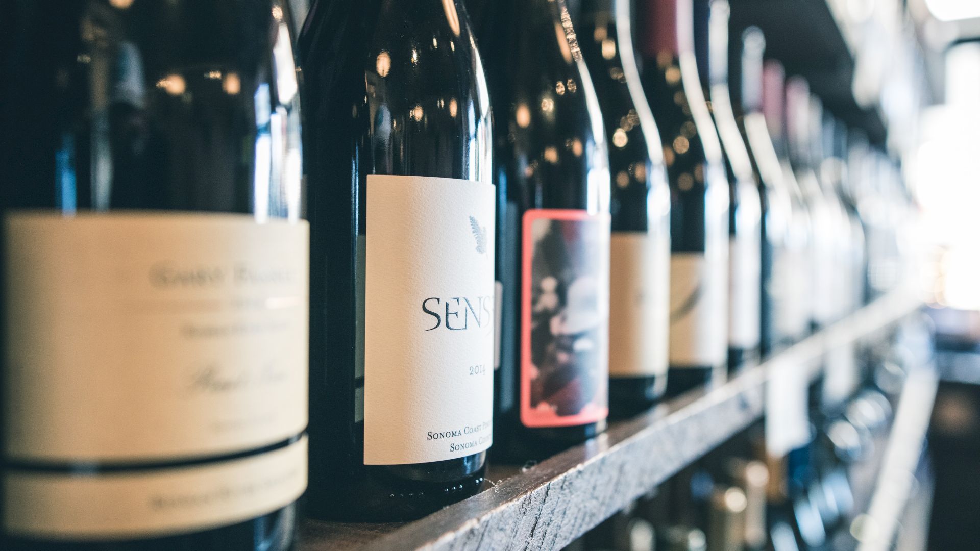 The image shows a row of wine bottles displayed on a shelf, with various labels and colors, located in what appears to be a wine store or a cellar.