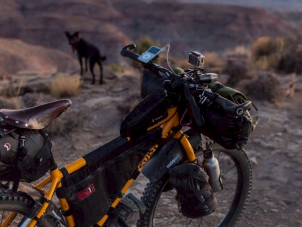 A loaded bike stands on a rocky terrain with a dog nearby, overlooking a vast canyon at sunset with colorful skies in the background.