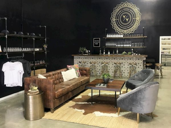 A stylish lounge area features a brown leather sofa, two grey chairs, a coffee table, and a decorative bar against a black wall with shelves.
