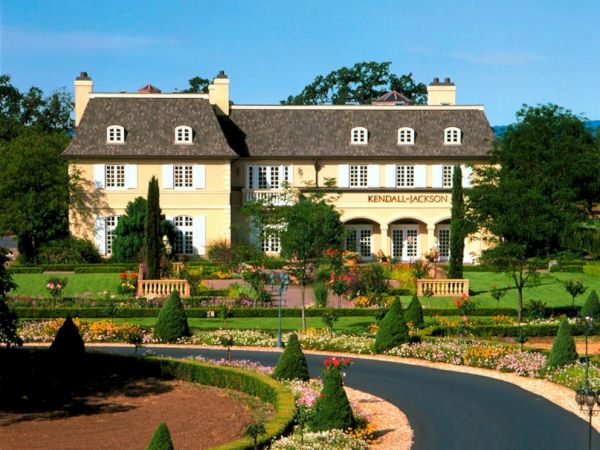 The image shows a grand house with a neatly manicured garden and a curved driveway leading up to the entrance, surrounded by lush greenery.