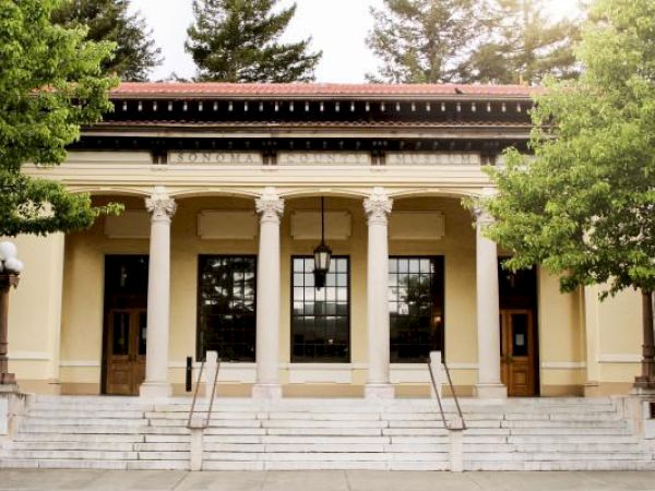 The image shows a building with four large columns, steps leading up to the entrance, flanked by two doors, with trees in the background.