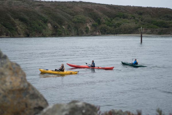 Three people are kayaking on a body of water with a hilly landscape in the background.
