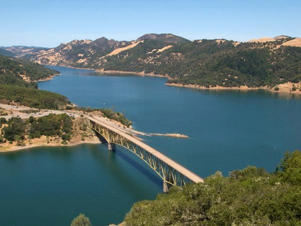 The image depicts a scenic view of a bridge spanning across a serene lake, surrounded by lush green hills and mountains under a clear blue sky.
