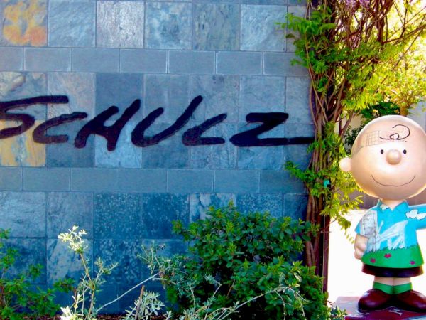 A statue of a smiling cartoon character stands next to a wall with "SCHULZ" written on it in large letters, surrounded by greenery and plants.