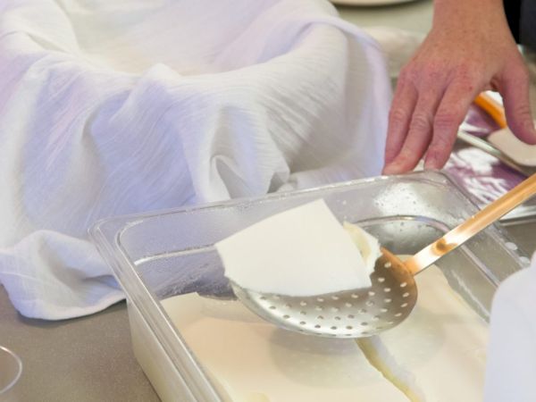 A person is preparing tofu, using a slotted spoon to lift a piece from a container, with a cloth-lined bowl in the background.