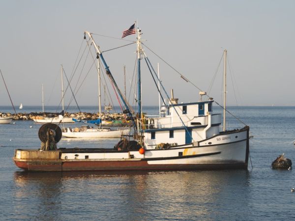 A fishing boat with an American flag docked in calm waters, alongside other smaller boats in the background, under a clear sky.