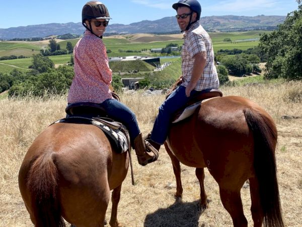 Two people wearing helmets and casual clothing are horseback riding in an open field with scenic hills and greenery in the background.