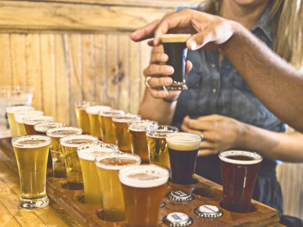 Several glasses of different types of beer on a wooden tray, with two people sampling them. Some bottle caps are visible on the table.