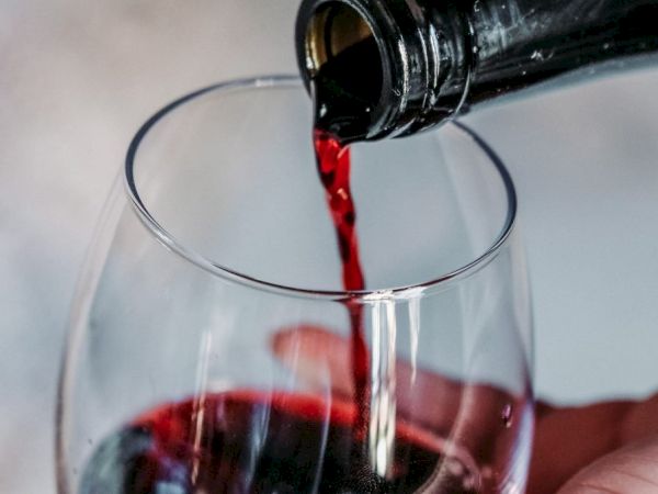 A bottle is pouring red wine into a glass, with a hand holding the glass, positioned against a blurred background.