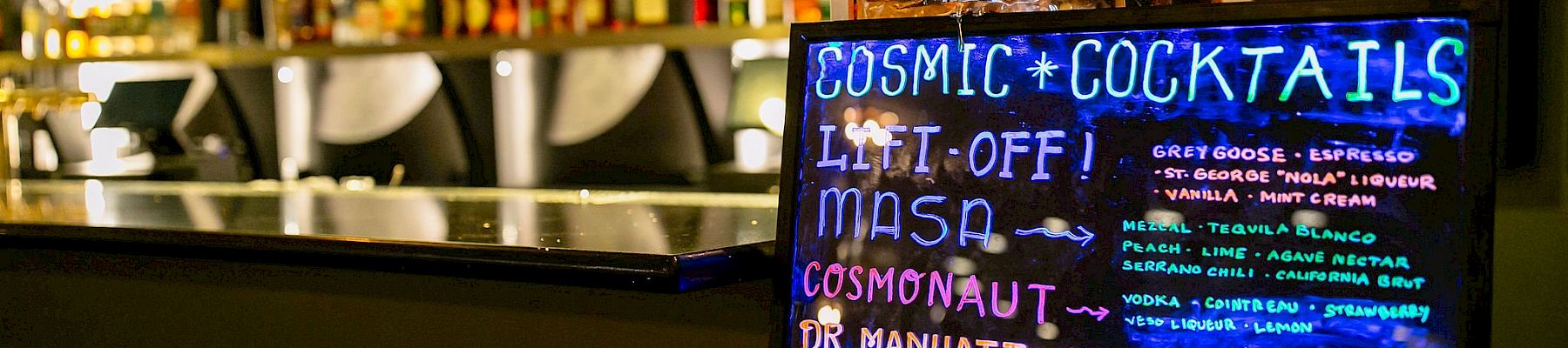 A neon sign at a bar advertises "Cosmic Cocktails" including drinks like Liftoff, Masa, Cosmonaut, and Dr. Manhattan. See you space cowboy.