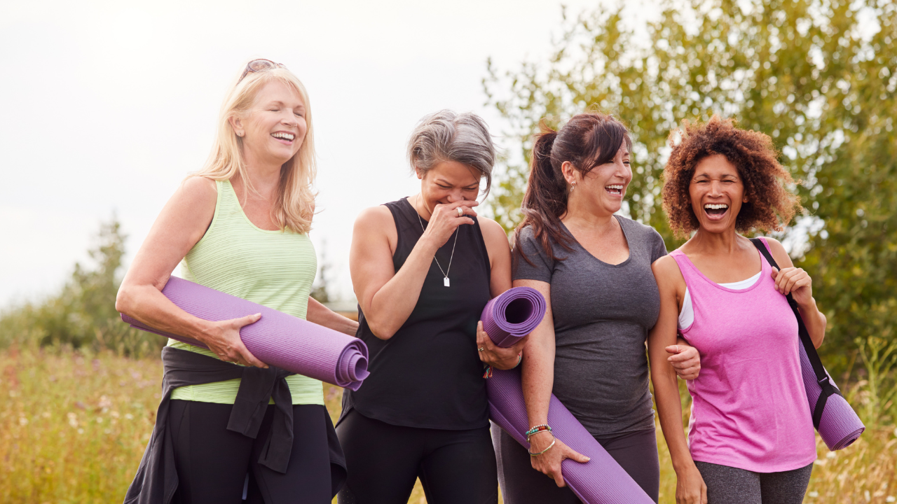 Four women are walking outdoors, laughing and chatting, each holding a yoga mat, dressed in workout clothing with a natural background.