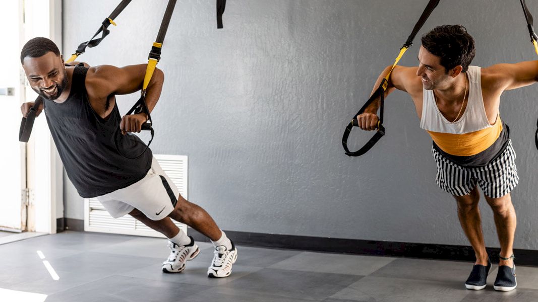 Two men are doing a suspension training exercise while smiling at each other in a gym. They are using TRX straps anchored to the ceiling.