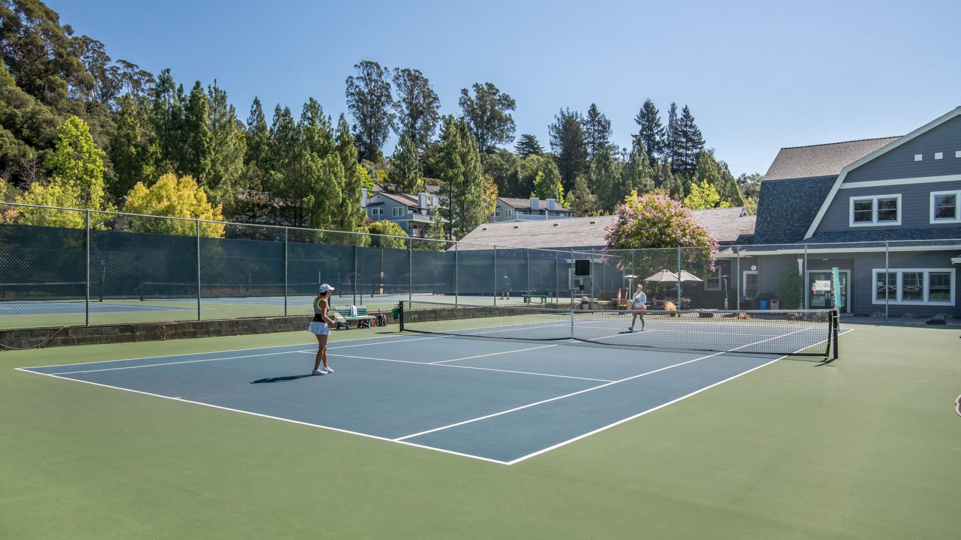 Two people are playing tennis on an outdoor court surrounded by trees and buildings.