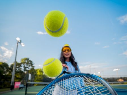 A person on a tennis court is hitting two tennis balls with a racket, with a blue sky in the background.