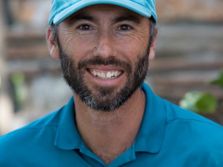 A man wearing a turquoise Nike polo and matching cap, smiling in front of a wooden background.