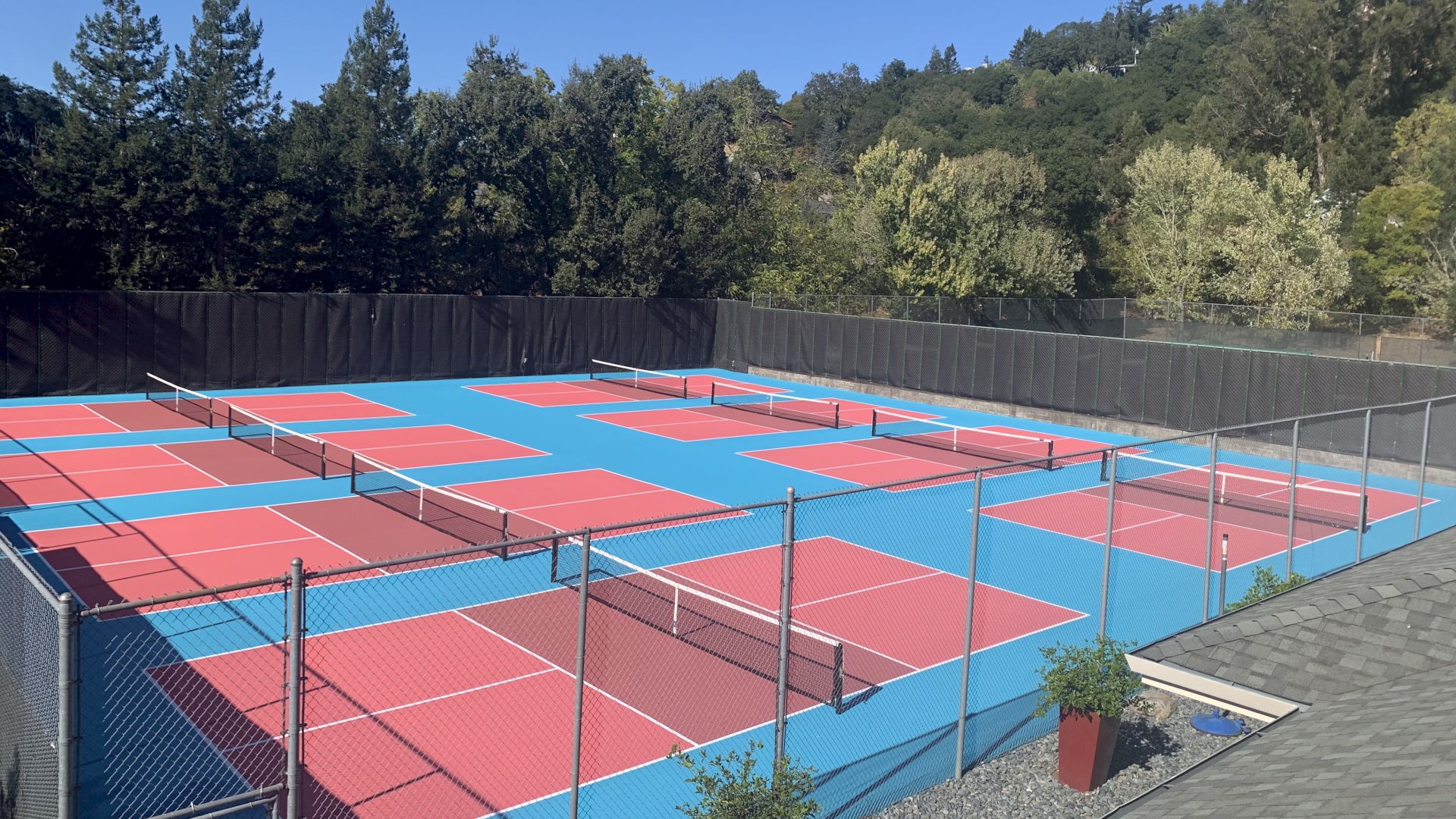 The image shows multiple pickleball courts with red surfaces and blue outlining, surrounded by fencing and trees in the background.