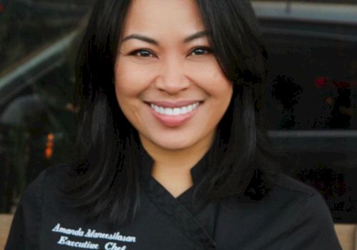 A person wearing a black chef's coat, smiling. The coat has a name and "Executive Chef" written on it.