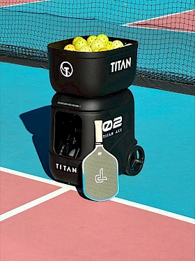 A black Titan ball hopper stacked on a white Titan ball machine, filled with yellow tennis balls, on a sunny outdoor court.