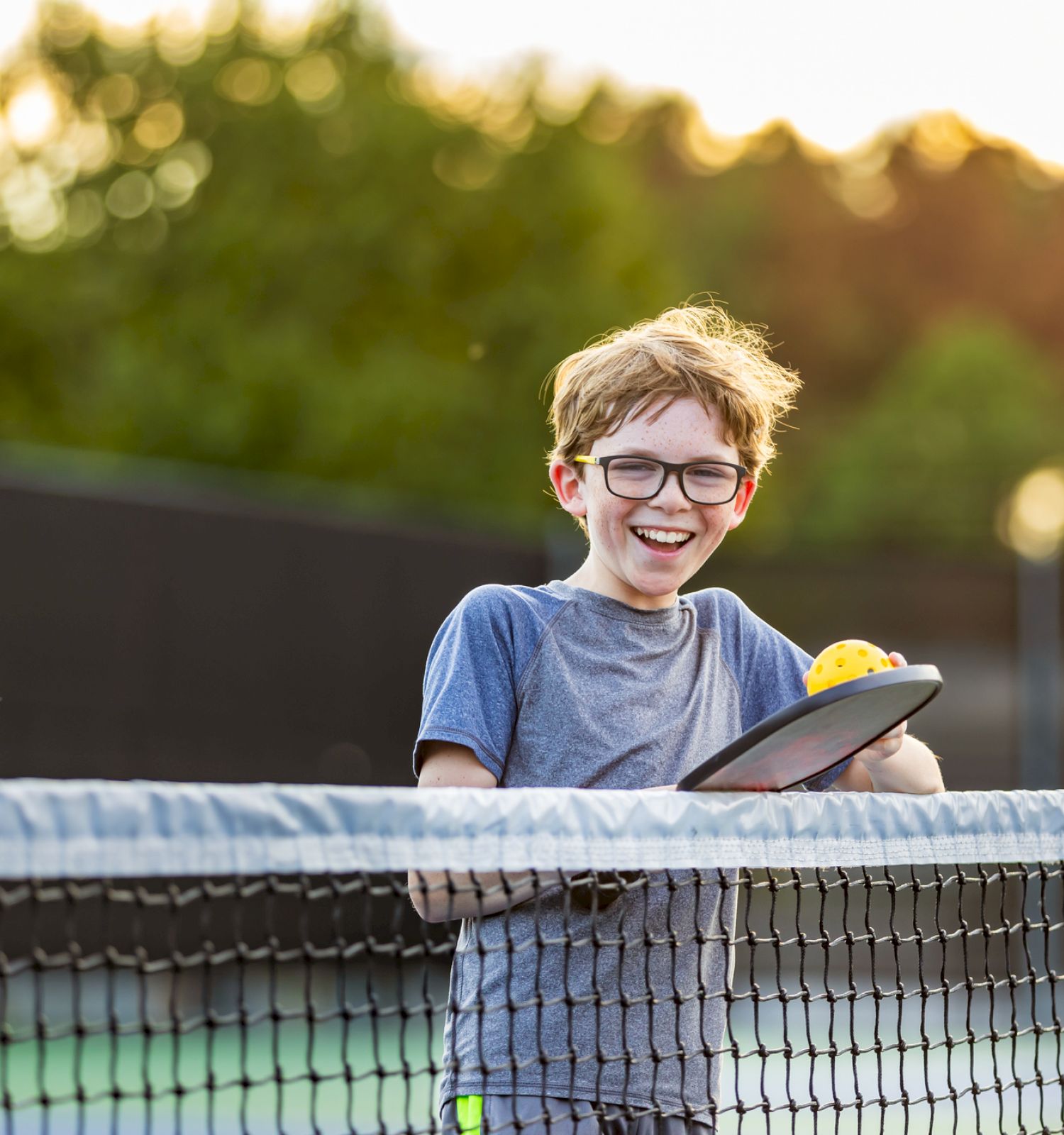 A boy stands at a tennis net, wearing glasses and a gray shirt, holding a tennis racket with a ball, smiling in an outdoor court.