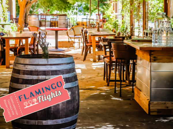 An outdoor seating area with wooden tables, chairs, barrel decor, and a bar. There's also a "FLAMINGO Flights" sign in the foreground.
