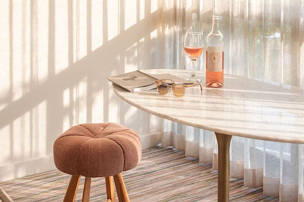 The image shows a cozy room with a round table, a pink stool, a bottle of rosé wine, and a filled glass near sheer curtains casting shadows.