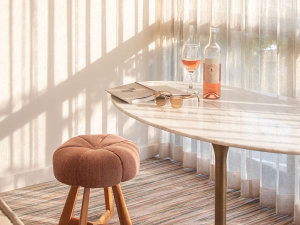 The image shows a cozy room with a round table, a pink stool, a bottle of rosé wine, and a filled glass near sheer curtains casting shadows.