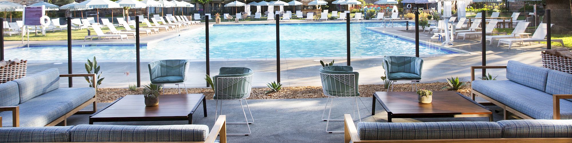 An outdoor seating area under a wooden pergola overlooks a clear swimming pool with lounge chairs and umbrellas surrounded by greenery and buildings.