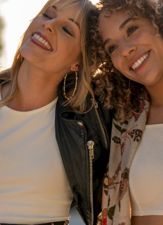 Two women are smiling and embracing each other outdoors, wearing casual clothing. Sunlight filters through the trees in the background.
