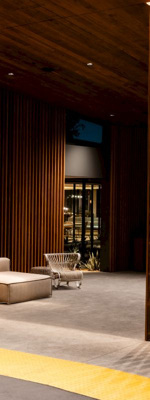 The image shows a modern, open-air lounge with wooden paneling, an overhead slatted ceiling, and a suspended chair, illuminated by warm lighting.
