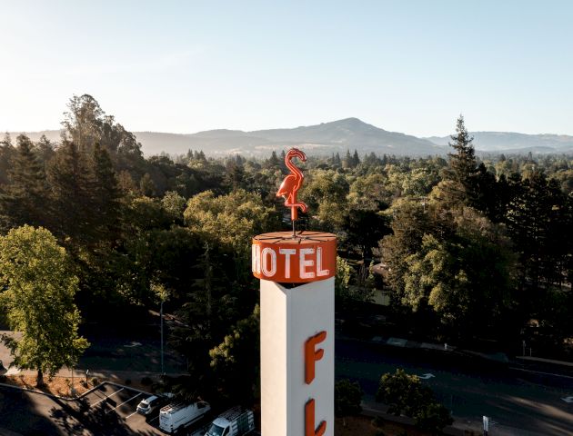 An image shows a tall Motel sign with a red flamingo on top, surrounded by trees, with a mountain range in the background and a few cars visible below.