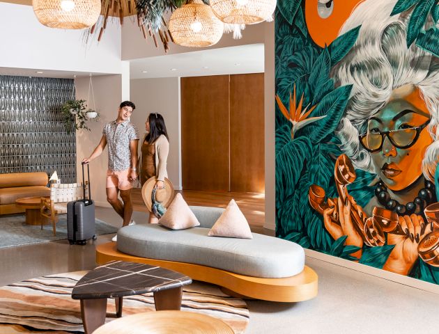 A couple with luggage stand in a modern lobby area with a large, colorful mural of a woman on the wall, wooden furniture, and hanging light fixtures.