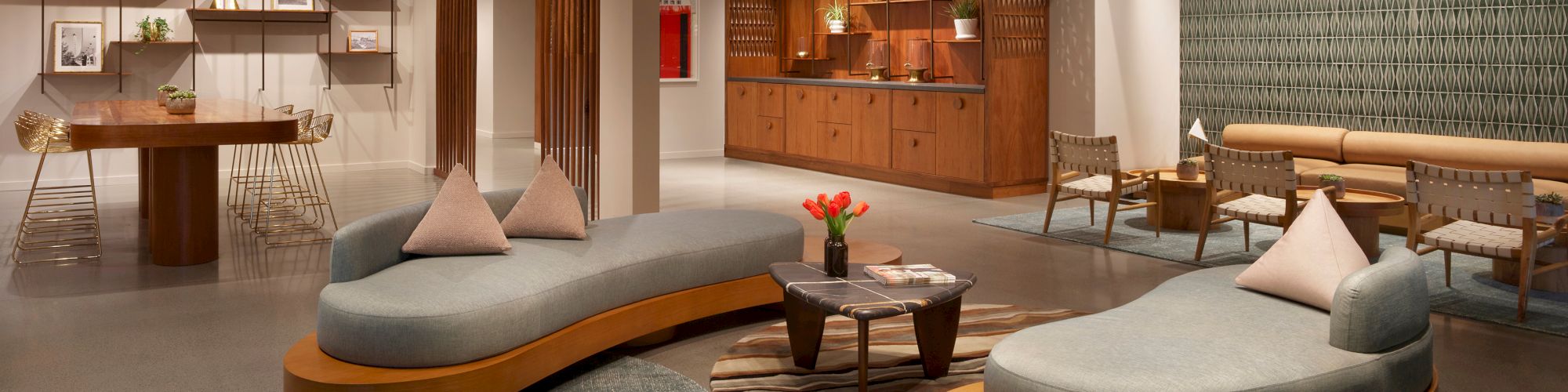 A modern, spacious lounge with stylish furniture, a kitchen area, a round table, and hanging lights. Decor includes potted plants and abstract art.