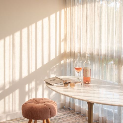 A cozy room with sunlight streaming in through sheer curtains, a small table with a drink and glass, and a round stool with wooden legs.