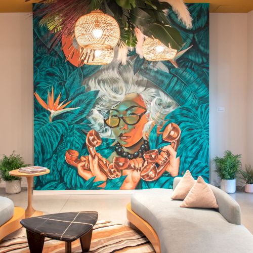 The image shows a vibrant mural of a woman with glasses, surrounded by plants and decor, in a modern room with sofas, a small table, and hanging lights.