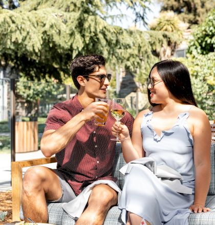 Two people are sitting outdoors, toasting drinks, and enjoying food on a table in a garden setting.