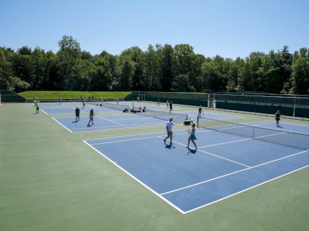The image shows an outdoor tennis facility with multiple courts and people playing tennis under a clear sky, surrounded by trees and greenery.