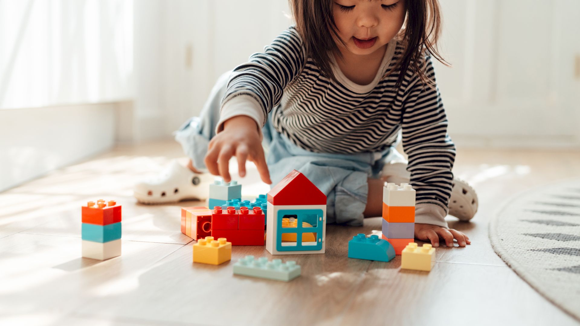 A child is playing on the floor with colorful toy building blocks in a sunlit room.