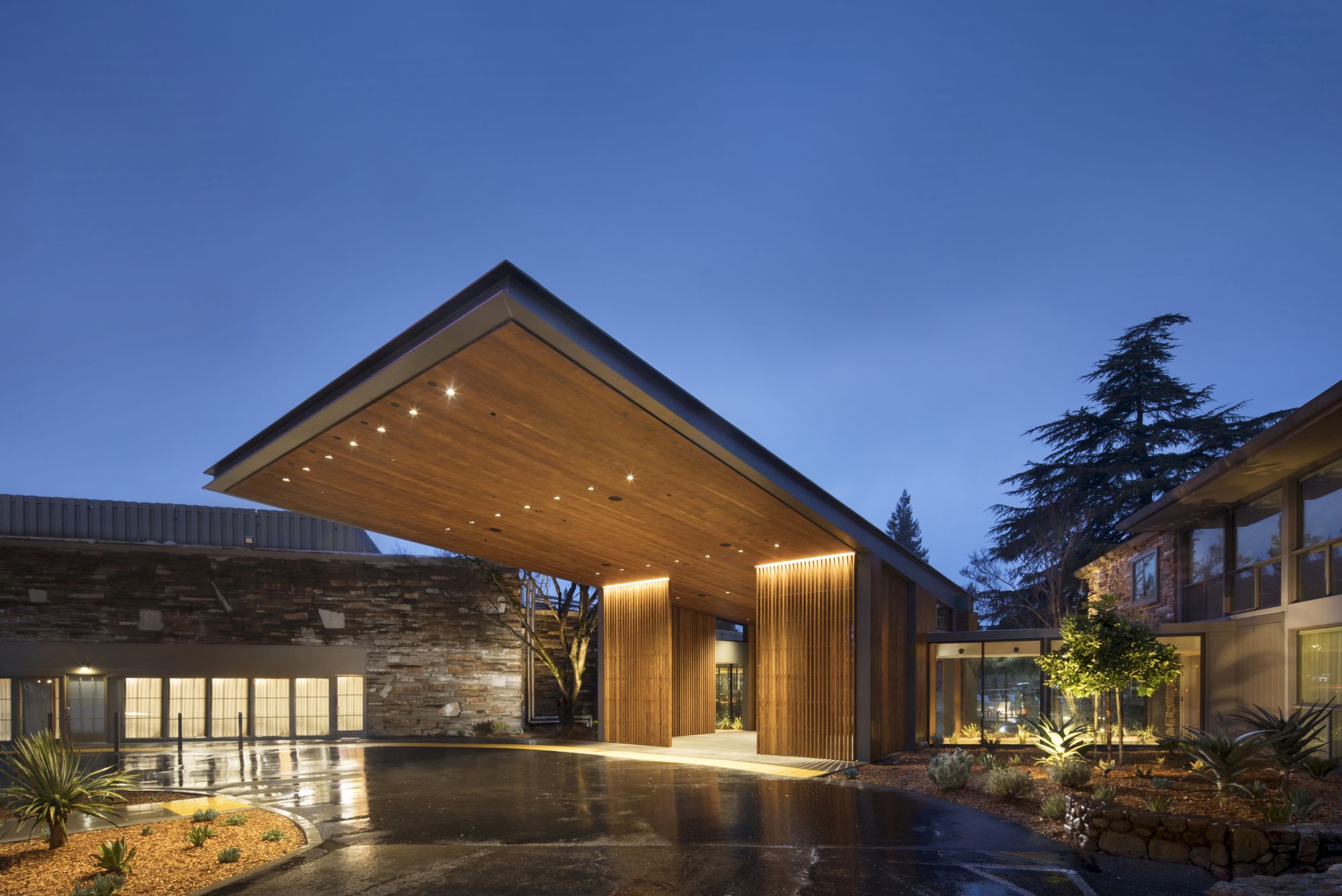 The image shows a modern building with a large wooden canopy entrance, surrounded by sleek landscaping and illuminated by lights at dusk.