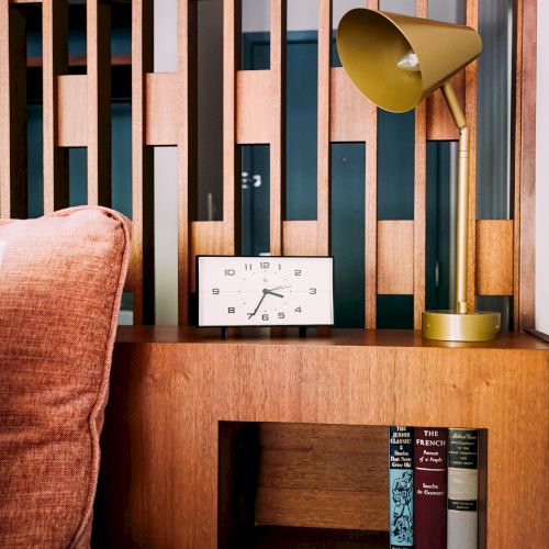 The image shows a bedside table with a modern gold lamp, a clock, and some books on a wooden rack. The background features a wooden panel design.