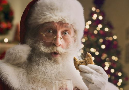 Santa Clause holding a glass of milk and a cookie, with a decorated Christmas tree in the background.