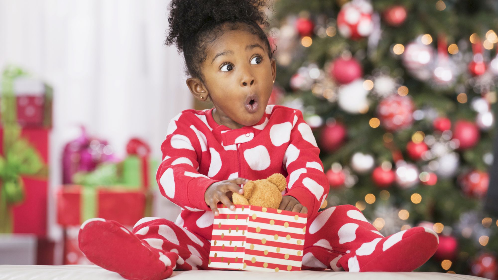 A child in red polka dot pajamas looks surprised, holding a gift, with a Christmas tree and presents in the background.