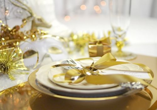 A festive table setting with gold accents, a mask, and a ribbon-wrapped gift against a blurred background with glowing lights.