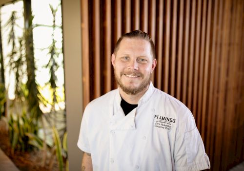 A person in a chef's uniform stands in a well-lit area with plants and wooden panels in the background, smiling at the camera.