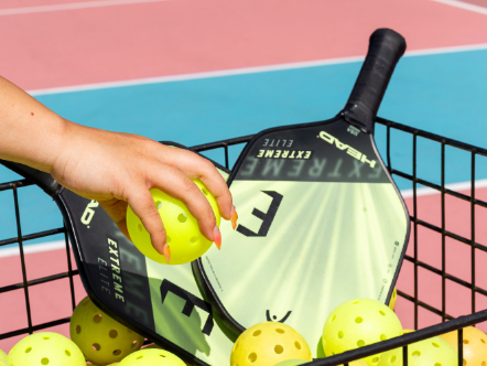 A hand places a bright yellow tennis ball into a basket with many other balls and a paddle resting among blue and pink tennis courts.