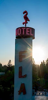A tall sign with "FLAMINGO HOTEL" and a flamingo figure, set against a sunrise or sunset sky, surrounded by trees and buildings.