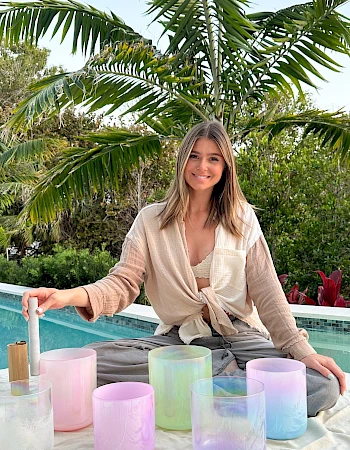 A smiling woman sits by a pool on a blanket, arranging pastel glass lanterns in pink, purple, and blue hues, with palm trees in the background.